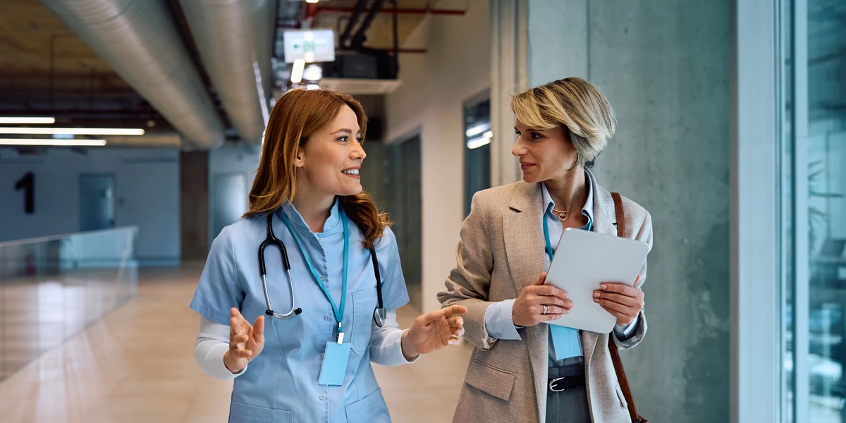 nurse and businesswoman talking while walking through hospital hallway.
